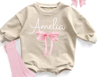 May include: A beige baby onesie with long sleeves and a pink bow on the front. The onesie has the name "Amelia" written in white script above the bow. The onesie is paired with pink socks and a pink bow hair clip.