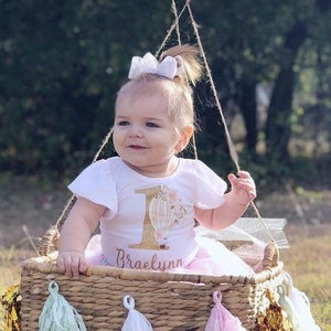May include: A baby girl wearing a white onesie with a hot air balloon graphic and the text "1 Braelyn" sits in a wicker basket decorated with pink, green, and gold tassels. A large pink balloon is attached to the basket.