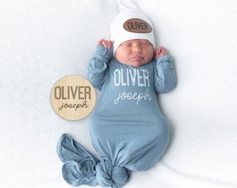 May include: A blue knotted baby gown with white embroidered text "Oliver Joseph" and a white knit hat with a brown leather name tag that says "Oliver". A wooden round sign with the text "Oliver Joseph" is on the white surface.