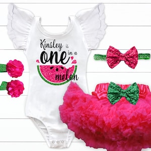 May include: A white bodysuit with a watermelon graphic and the text "Kinsley is one in a melon". The bodysuit has a pink bow headband, two pink flower hair clips, and a pink tulle skirt with a green bow.