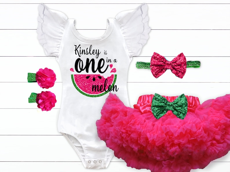 May include: A white bodysuit with a watermelon graphic and the text "Kinsley is one in a melon". The bodysuit has a pink bow headband, two pink flower hair clips, and a pink tulle skirt with a green bow.