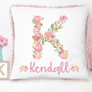 Op de afbeelding: Een wit kussen met een roze bloemige letter K en de naam "Kendall" in roze schrift. Het kussen heeft een roze pluche rand.