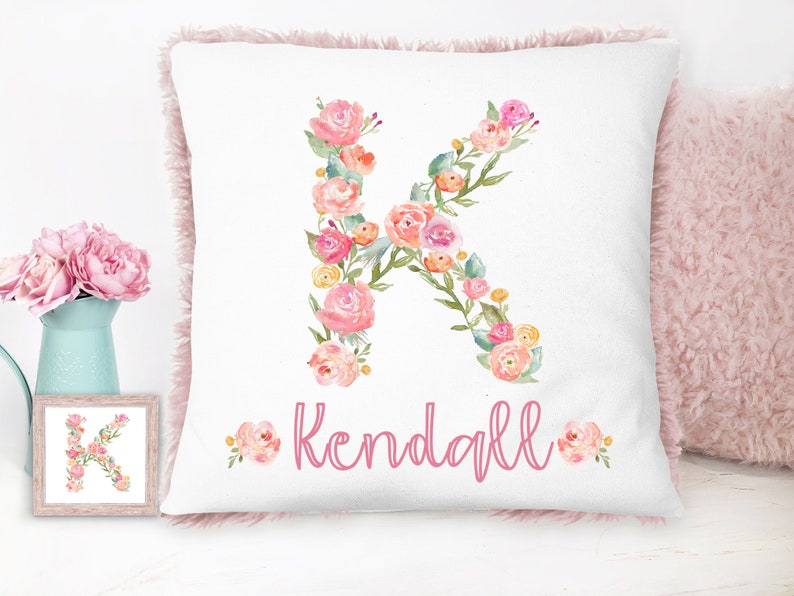 Op de afbeelding: Een wit kussen met een roze bloemige letter K en de naam "Kendall" in roze schrift. Het kussen heeft een roze pluche rand.