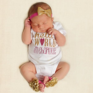 Puede incluir: Un bebé recién nacido con un body blanco con letras doradas brillantes que dicen "hello world i'm charlotte" y una diadema rosa con un lazo dorado. El bebé lleva botines de tobillo de flores doradas y rosas.