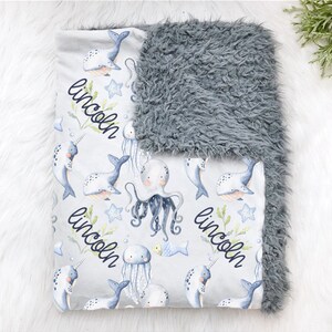 May include: A gray and white baby blanket with a plush gray side and a white side with a repeating pattern of narwhals, jellyfish, and octopuses. The blanket is personalized with the name "Lincoln" in a black cursive font.