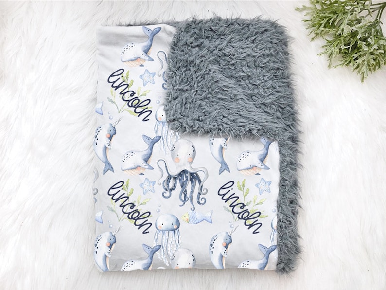 May include: A gray and white baby blanket with a plush gray side and a white side with a repeating pattern of narwhals, jellyfish, and octopuses. The blanket is personalized with the name "Lincoln" in a black cursive font.