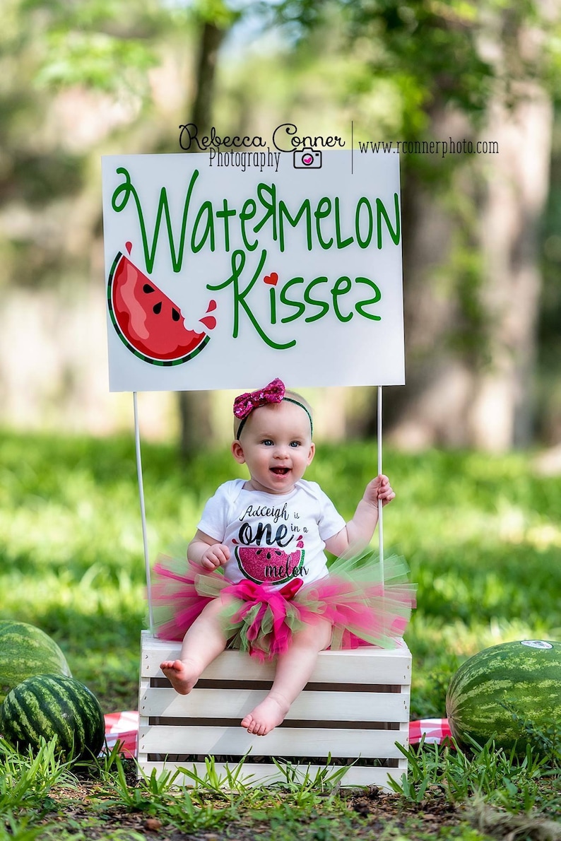 May include: A baby girl wearing a white onesie with a watermelon graphic and the text "Aisceigh is one in a melon" sits on a white wooden crate. She is wearing a pink and green tutu and a pink headband with glitter. There is a sign behind her that says "Watermelon Kisses" with a watermelon graphic. There are two watermelons on the ground.