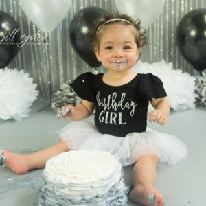 May include: A baby girl wearing a black shirt with silver glitter lettering that says "birthday girl" and a white tulle tutu sits in front of a cake with white and gray frosting. The baby is covered in frosting and has a happy expression on her face.