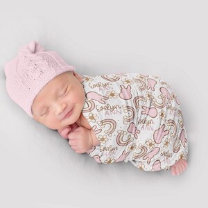 Puede incluir: Una manta de arrullo rosa y blanca con un patr&oacute;n repetido de arco&iacute;ris, conejos y flores. La manta est&aacute; personalizada con el nombre "Evelyn Ann".