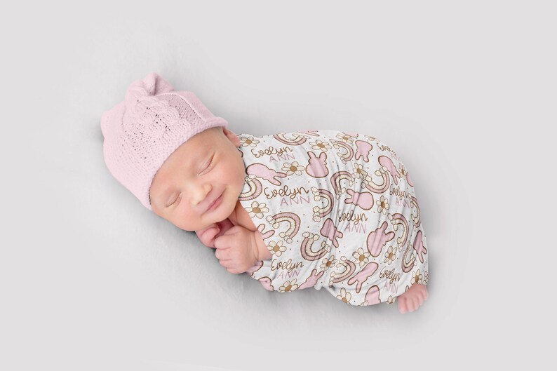 Puede incluir: Una manta de arrullo rosa y blanca con un patr&oacute;n repetido de arco&iacute;ris, conejos y flores. La manta est&aacute; personalizada con el nombre "Evelyn Ann".