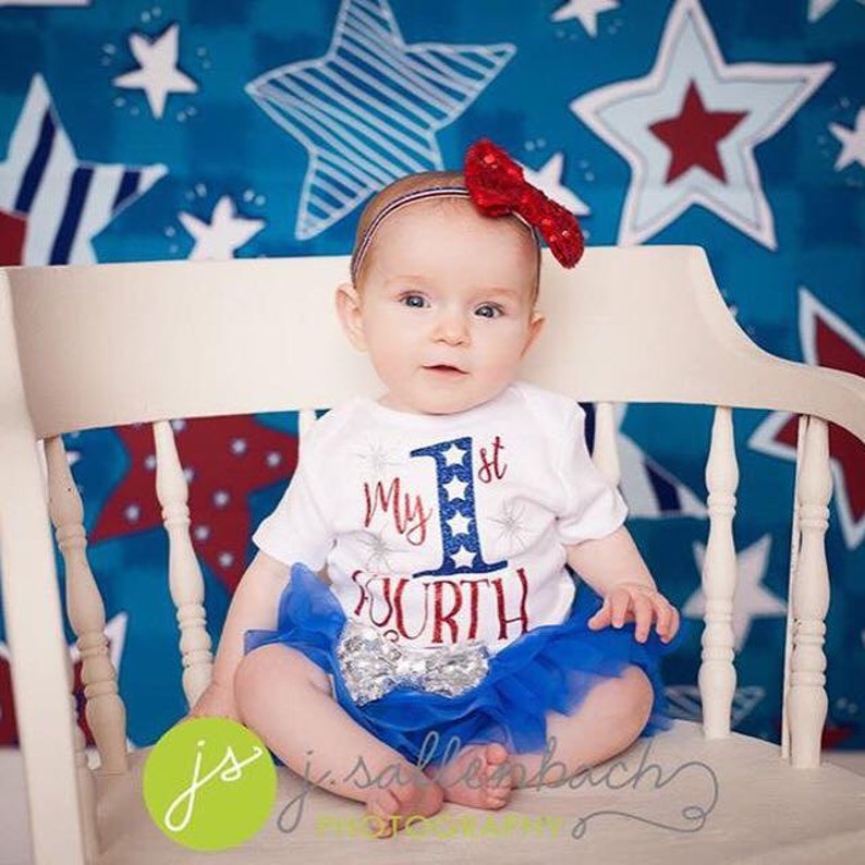 Puede incluir: Una ni&ntilde;a peque&ntilde;a con un body blanco con el texto "My 1st Fourth" en rojo, blanco y azul. Lleva una falda de tul azul con lentejuelas plateadas y una diadema roja con lazo. La beb&eacute; est&aacute; sentada en una silla blanca frente a un fondo azul y rojo con estampado de estrellas.