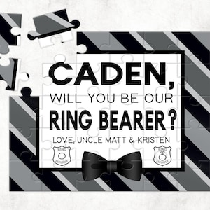 May include: A personalized puzzle with a black and white striped background. The puzzle asks "CADEN, WILL YOU BE OUR RING BEARER?" with the names "LOVE, UNCLE MATT & KRISTEN" below. Includes a black bow tie graphic.