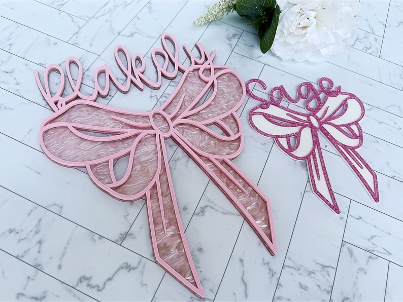 May include: Two pink wooden signs with the names "Blakeley" and "Sage" written in cursive. Each sign features a pink bow with a white center.
