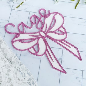 May include: A pink glitter bow with the word "Sage" written in cursive on top. The bow is made of white cardstock and has a pink glitter outline.