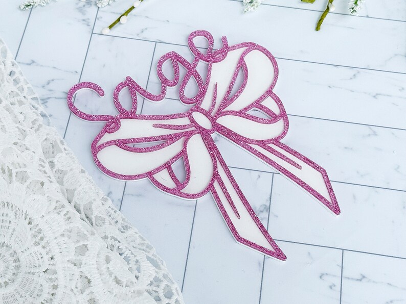 May include: A pink glitter bow with the word "Sage" written in cursive on top. The bow is made of white cardstock and has a pink glitter outline.