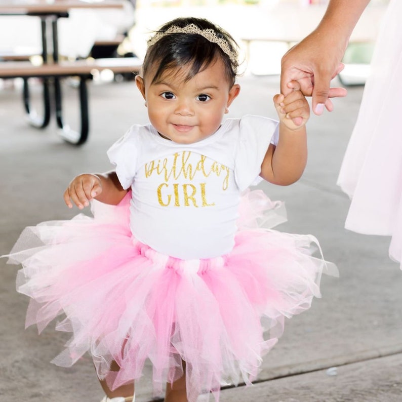May include: A baby girl wearing a white onesie with gold glitter lettering that says "birthday girl" and a pink tulle tutu. She is holding hands with an adult and smiling.