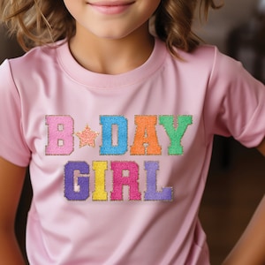 Puede incluir: Camiseta rosa de manga corta con el texto "B-DAY GIRL" en colores arcoíris con detalles de purpurina dorada.