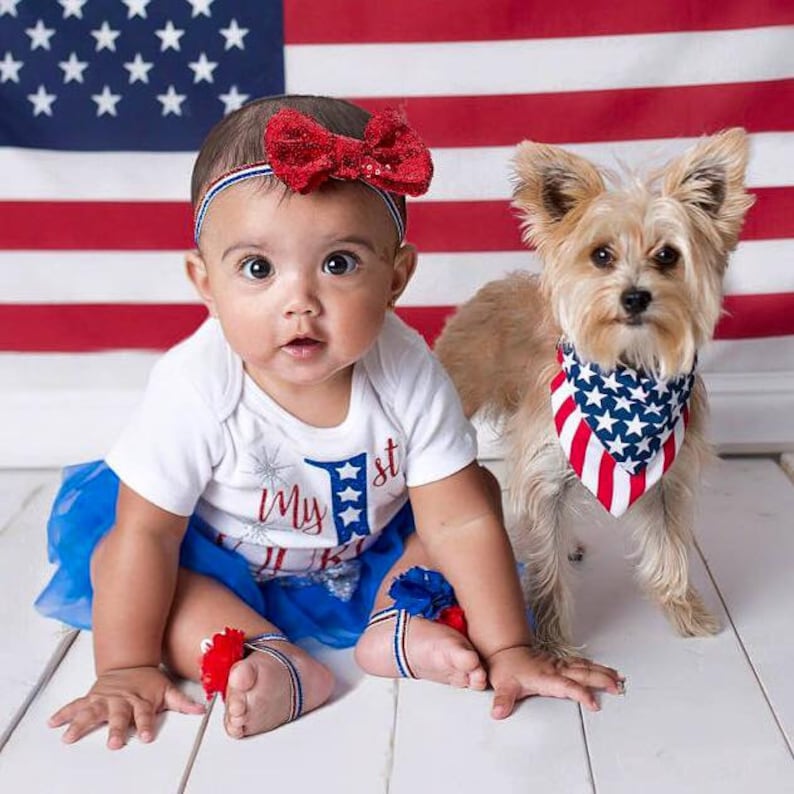 Puede incluir: Una ni&ntilde;a peque&ntilde;a con un body blanco con el texto "My 1st" y una falda de tul azul, una diadema roja con lazo y tobilleras rojas, blancas y azules. Un perro peque&ntilde;o con una bandana roja, blanca y azul est&aacute; sentado junto a la beb&eacute;. Una bandera estadounidense est&aacute; en el fondo.