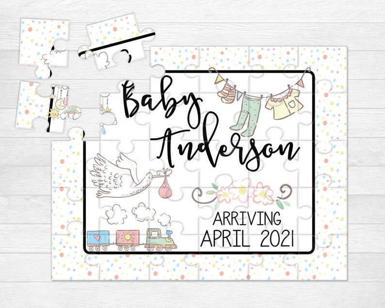 May include: A white jigsaw puzzle with a colorful confetti pattern. The puzzle features a stork carrying a baby, a train, and a clothesline with baby clothes. The text on the puzzle reads "Baby Anderson Arriving April 2021".