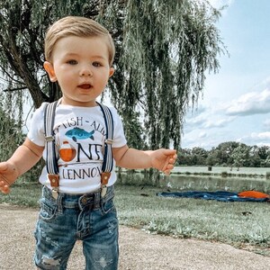 Puede incluir: Un ni&ntilde;o peque&ntilde;o con una camiseta blanca con un gr&aacute;fico de un pez azul y naranja y el texto "Fish-All One Lennox". Tambi&eacute;n lleva pantalones cortos de mezclilla azul y tirantes.