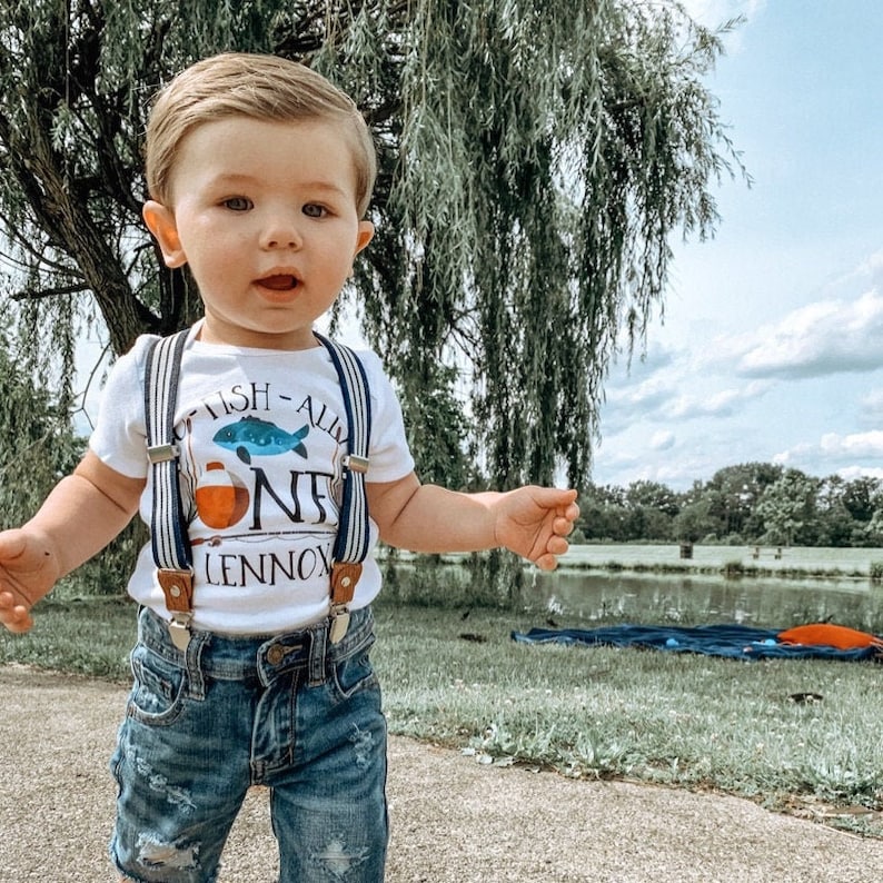 Puede incluir: Un ni&ntilde;o peque&ntilde;o con una camiseta blanca con un gr&aacute;fico de un pez azul y naranja y el texto "Fish-All One Lennox". Tambi&eacute;n lleva pantalones cortos de mezclilla azul y tirantes.