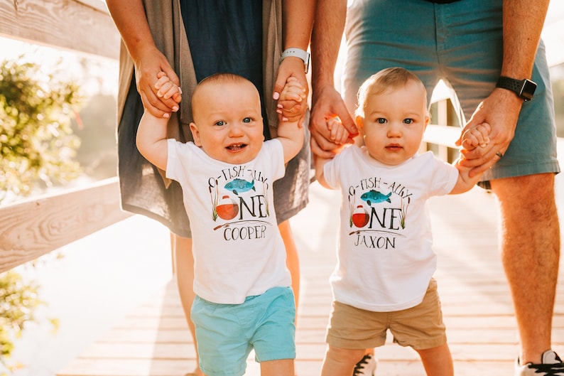Puede incluir: Dos beb&eacute;s varones con camisetas blancas con el texto "G-Fish-All-Y'all One" y sus nombres, Cooper y Jaxon. Est&aacute;n cogidos de la mano de sus padres y sonriendo.