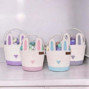 May include: Three pastel colored Easter baskets with bunny ears. The baskets are made of woven rope and have a white base with a colored trim. The baskets are filled with colorful Easter eggs.