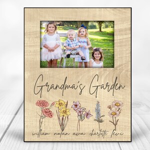 May include: A wooden picture frame with a light brown background. The frame has a white mat and a picture of four children in the center. The text "Grandma's Garden" is printed below the picture. There are five flowers with names below them: William, Nolan, Ava, Charlotte, Levi.