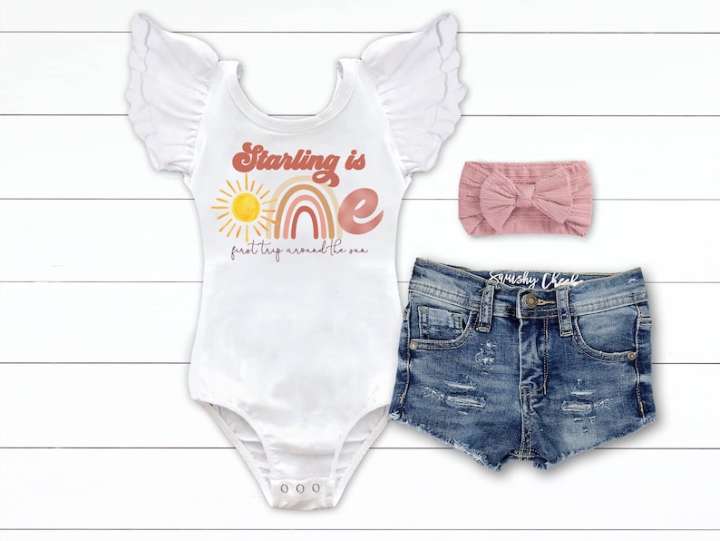Pode incluir: Um body branco com um arco-&iacute;ris e um sol rosa, laranja e amarelo. O texto "Starling is one first trip around the sun" est&aacute; impresso no body. Tamb&eacute;m est&aacute; inclu&iacute;do uma faixa de cabe&ccedil;a rosa e cal&ccedil;&otilde;es de ganga.