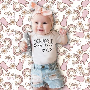 Puede incluir: Un beb&eacute; con un body blanco con el texto "SNUGGLE BUNNY" y una diadema rosa est&aacute; sentado en una manta blanca con un patr&oacute;n de conejito rosa y marr&oacute;n y arco&iacute;ris. La manta tiene el nombre "Evelyn Ann" repetido en todo el patr&oacute;n.