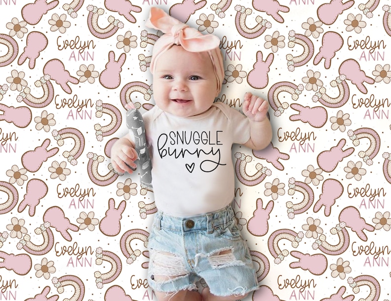 Puede incluir: Un beb&eacute; con un body blanco con el texto "SNUGGLE BUNNY" y una diadema rosa est&aacute; sentado en una manta blanca con un patr&oacute;n de conejito rosa y marr&oacute;n y arco&iacute;ris. La manta tiene el nombre "Evelyn Ann" repetido en todo el patr&oacute;n.