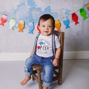 Puede incluir: Un beb&eacute; var&oacute;n con una camisa blanca con tirantes azules y un gr&aacute;fico de pez azul y rojo que dice "I Fish-ally One Christian". Est&aacute; sentado en una silla de madera con un atuendo de mezclilla azul.