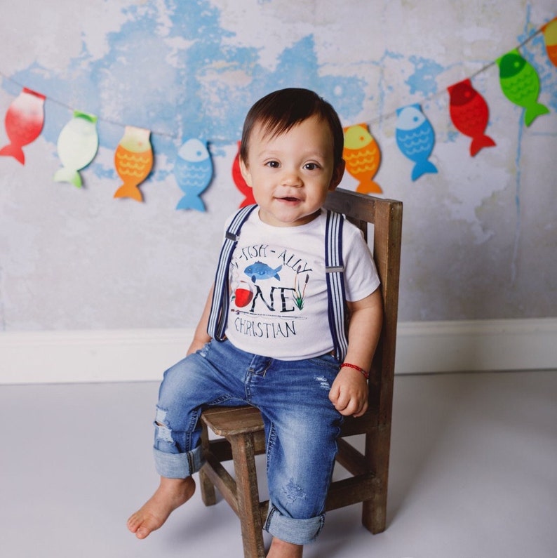 Puede incluir: Un beb&eacute; var&oacute;n con una camisa blanca con tirantes azules y un gr&aacute;fico de pez azul y rojo que dice "I Fish-ally One Christian". Est&aacute; sentado en una silla de madera con un atuendo de mezclilla azul.