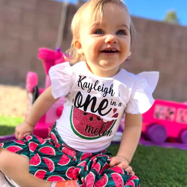 Puede incluir: Una ni&ntilde;a peque&ntilde;a con una camiseta blanca con un gr&aacute;fico de sand&iacute;a y el texto "Kayleigh is one in a melon". Tambi&eacute;n lleva una falda de estampado de sand&iacute;a verde y rosa.