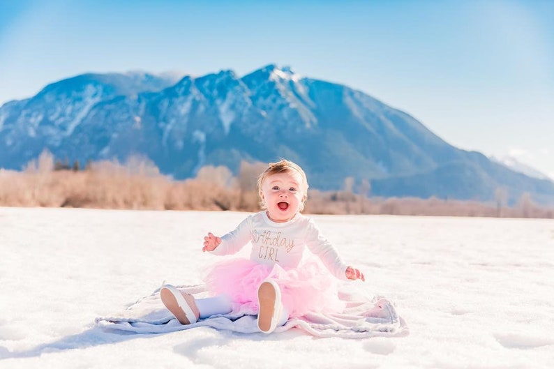 May include: A baby girl wearing a white long-sleeved shirt with the text "1st Birthday Girl" in gold glitter, a pink tulle tutu, and white shoes, sitting on a white blanket in the snow with a mountain range in the background.