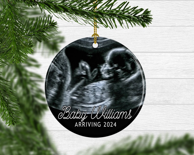 May include: A round, white ceramic ornament with a black and white ultrasound image of a baby. The text "Baby Williams Arriving 2024" is printed in white on the ornament.