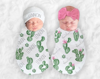 May include: Two baby swaddles with cactus print. One swaddle is white with green cacti and the name Lennon repeated on it. The other swaddle is white with green cacti and pink flowers and the name Amelia repeated on it. A white hat with the name Lennon on it is on the baby in the first swaddle. A pink headband is on the baby in the second swaddle.