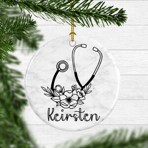 May include: White ceramic Christmas ornament with a black and white illustration of a stethoscope and flowers. The ornament has a marble-like background and the name "Keirsten" is written below the illustration.