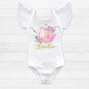 Puede incluir: Body blanco con mangas con volantes, con una tetera rosa con detalles florales y el n&uacute;mero "2" en dorado. El texto "Laila" est&aacute; escrito en dorado debajo de la tetera.