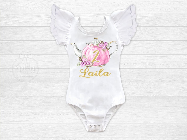 Puede incluir: Body blanco con mangas con volantes, con una tetera rosa con detalles florales y el n&uacute;mero "2" en dorado. El texto "Laila" est&aacute; escrito en dorado debajo de la tetera.