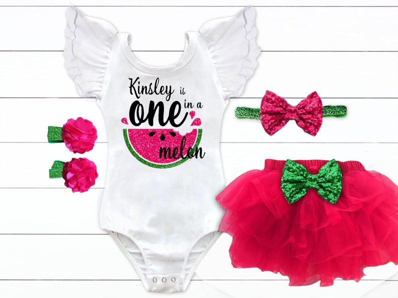 Puede incluir: Un body blanco con un gr&aacute;fico de sand&iacute;a y el texto "Kinsley is one in a melon". El body tiene detalles de purpurina rosa y verde. El conjunto tambi&eacute;n incluye una falda de tul rosa con un lazo de purpurina verde, una diadema con un lazo de purpurina rosa y dos pinzas para el pelo de flores rosas.