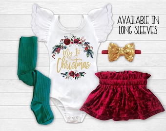 May include: A white bodysuit with a gold glitter 'My 1st Christmas' design and a red and green floral wreath. The bodysuit has ruffled sleeves. The outfit also includes a red velvet skirt with a ruffled hem and a gold bow headband.