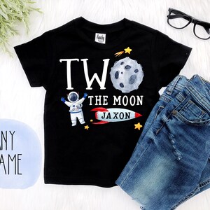 May include: Black short-sleeved t-shirt with a white astronaut graphic, a moon, a rocket, and the text "Two the Moon Jaxon".
