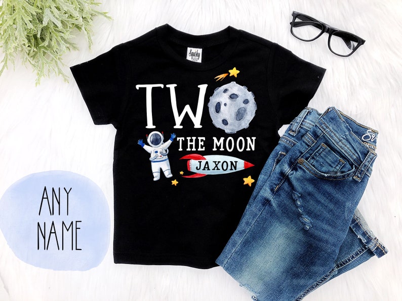 May include: Black short-sleeved t-shirt with a white astronaut graphic, a moon, a rocket, and the text "Two the Moon Jaxon".