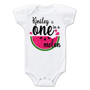 Puede incluir: Un body blanco para beb&eacute; con un gr&aacute;fico de sand&iacute;a rosa y verde y el texto "Kinsley is one in a melon".