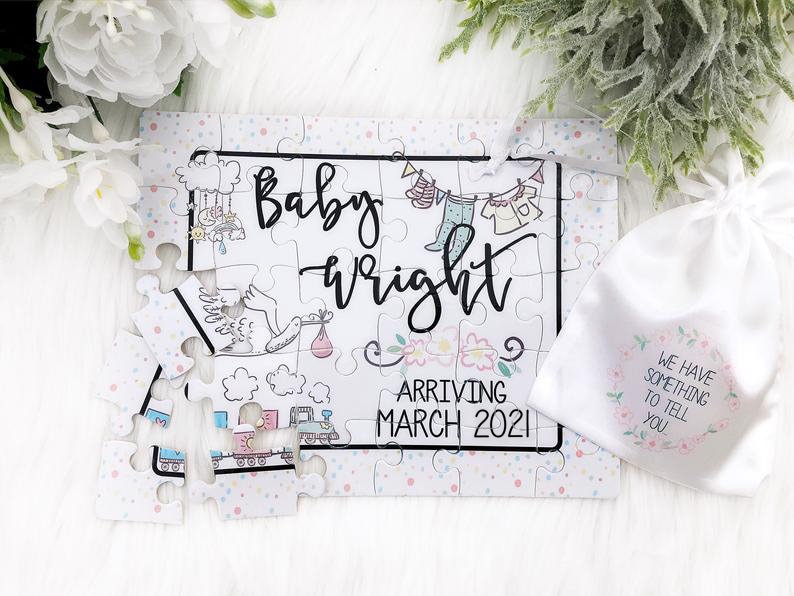 May include: A white puzzle with a pink and blue confetti background. The puzzle is assembled to reveal the text "Baby Wright Arriving March 2021". The puzzle is accompanied by a white drawstring bag with the text "We have something to tell you" printed on it.