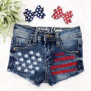 May include: Blue denim shorts with a distressed hem and a white star and red stripe American flag design. The shorts have a "Smushy Cheeks" label on the back.
