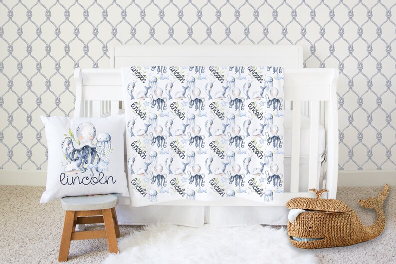 May include: A white baby blanket with a nautical theme featuring an octopus, jellyfish, and the name "Lincoln" repeated throughout the design. The blanket is displayed on a white crib with a gray rope pattern wallpaper.