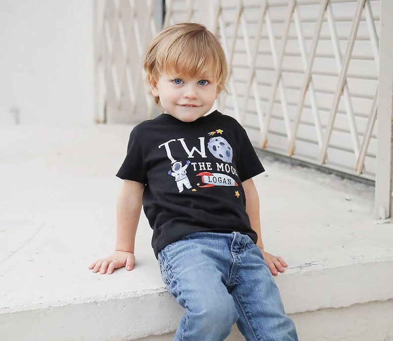 May include: A young boy wearing a black t-shirt with a graphic of an astronaut, a moon, and the text "Two the Moon Logan". He is wearing blue jeans and sitting on a concrete step.