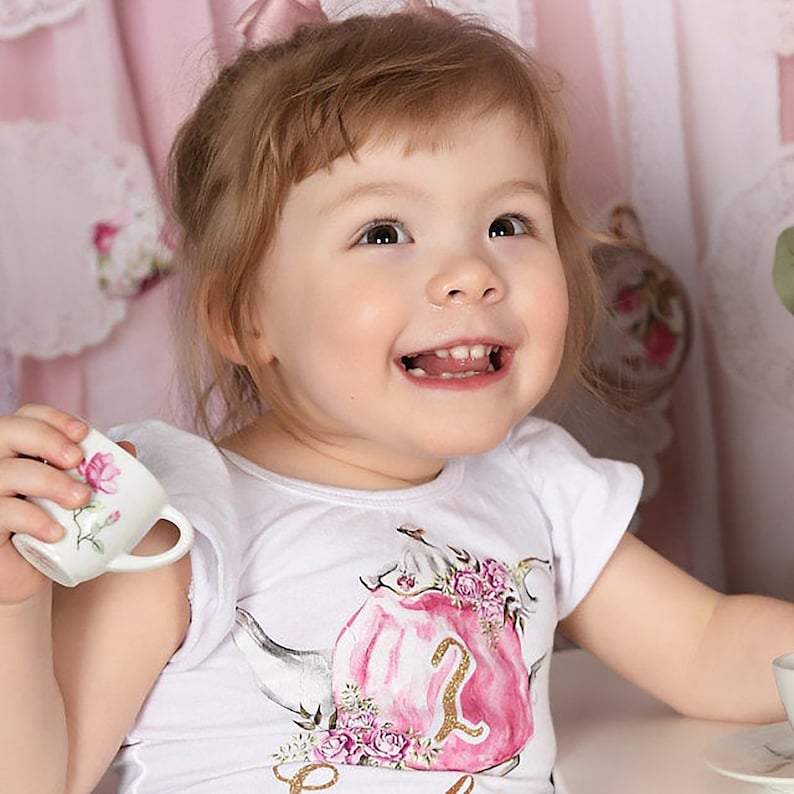 Puede incluir: Una ni&ntilde;a peque&ntilde;a con una camiseta blanca con un dise&ntilde;o de tetera rosa y flores y el n&uacute;mero "2" en purpurina dorada. Est&aacute; sosteniendo una peque&ntilde;a taza de t&eacute; blanca con flores rosas.